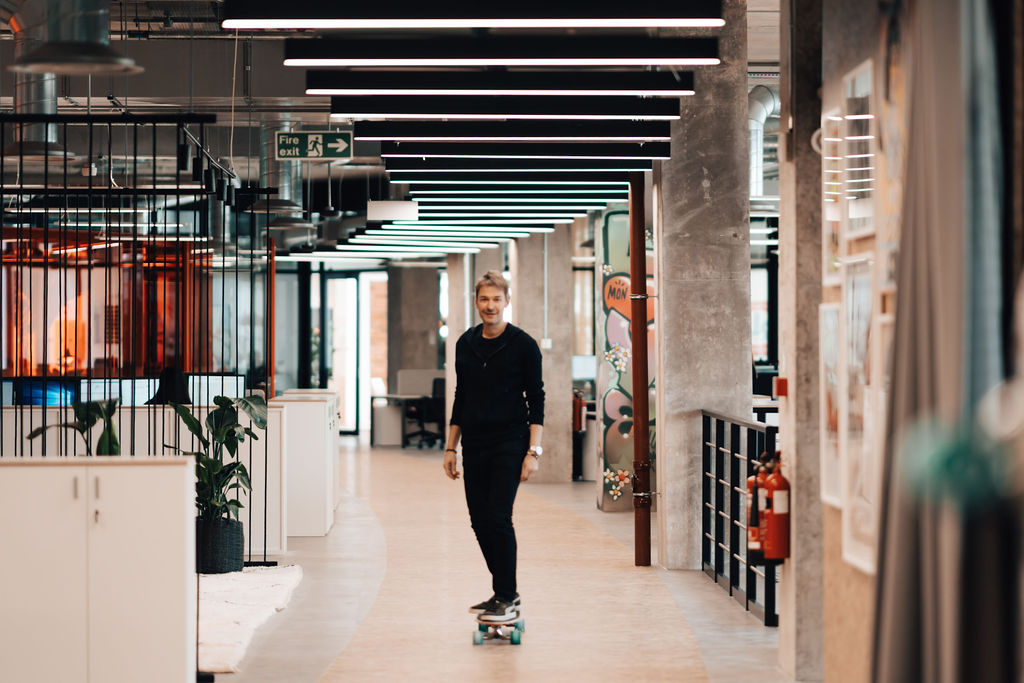 A modern office hallway designed by Modus Workspace for SLG, featuring industrial elements like exposed concrete pillars and a grid of ceiling lights. A person is casually riding a skateboard through the space, reflecting a relaxed and creative work environment. The hallway is lined with various office elements, including plants and wall decor, contributing to the stylish and dynamic atmosphere.