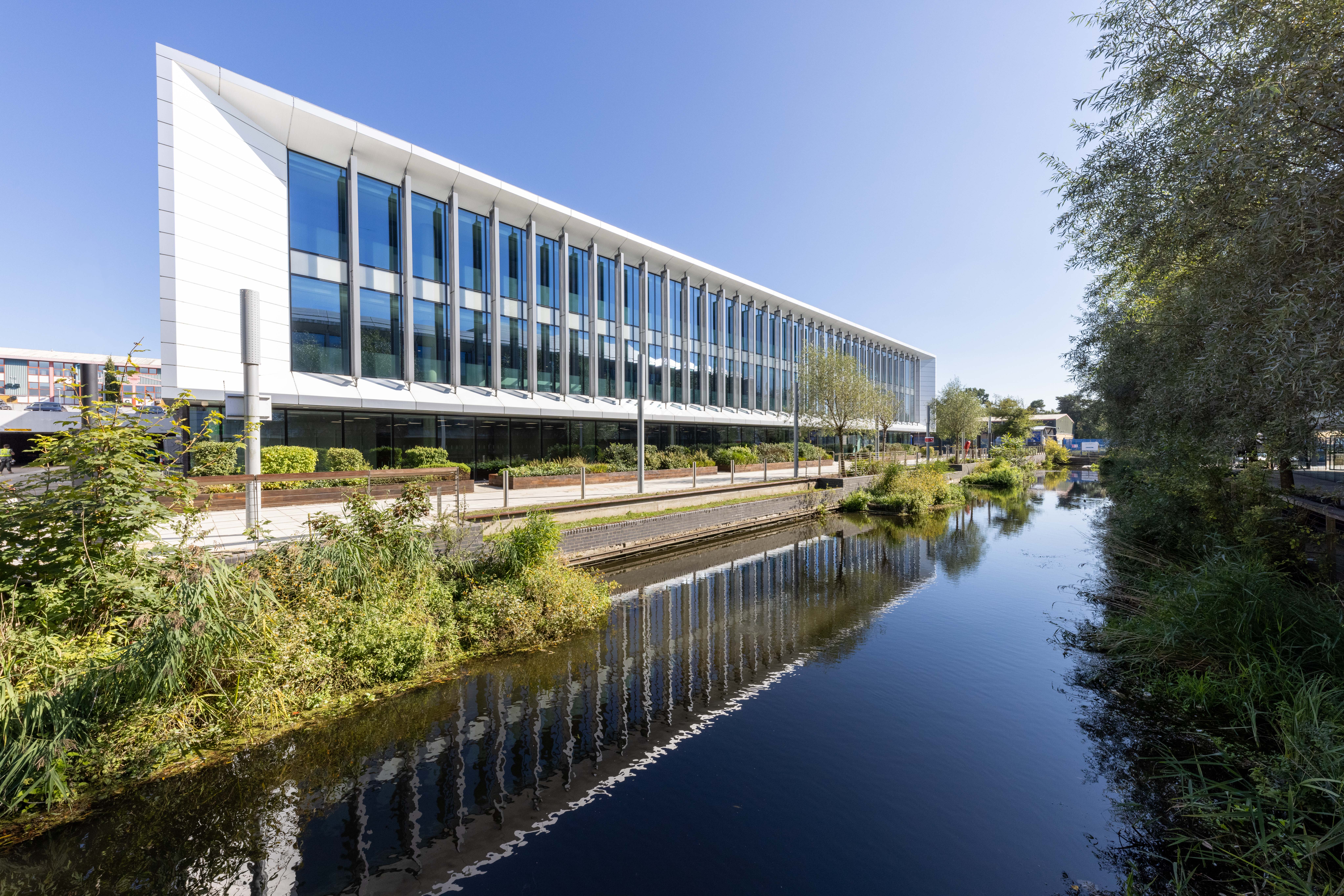 The Dock office building with a modern glass facade is reflected in a nearby canal under a clear blue sky.