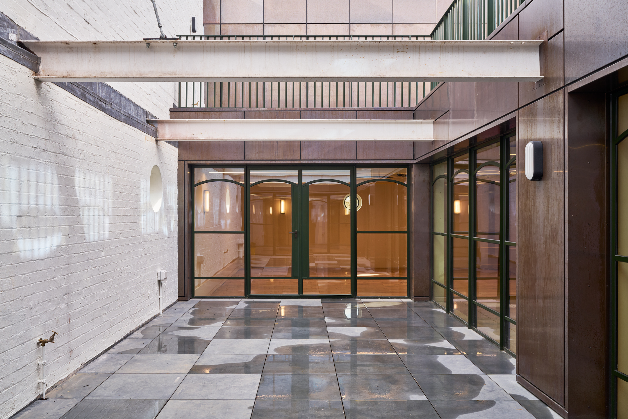 A clean, sleek courtyard space with glass doors framed by metal and dark green structures, leading into a modern indoor are