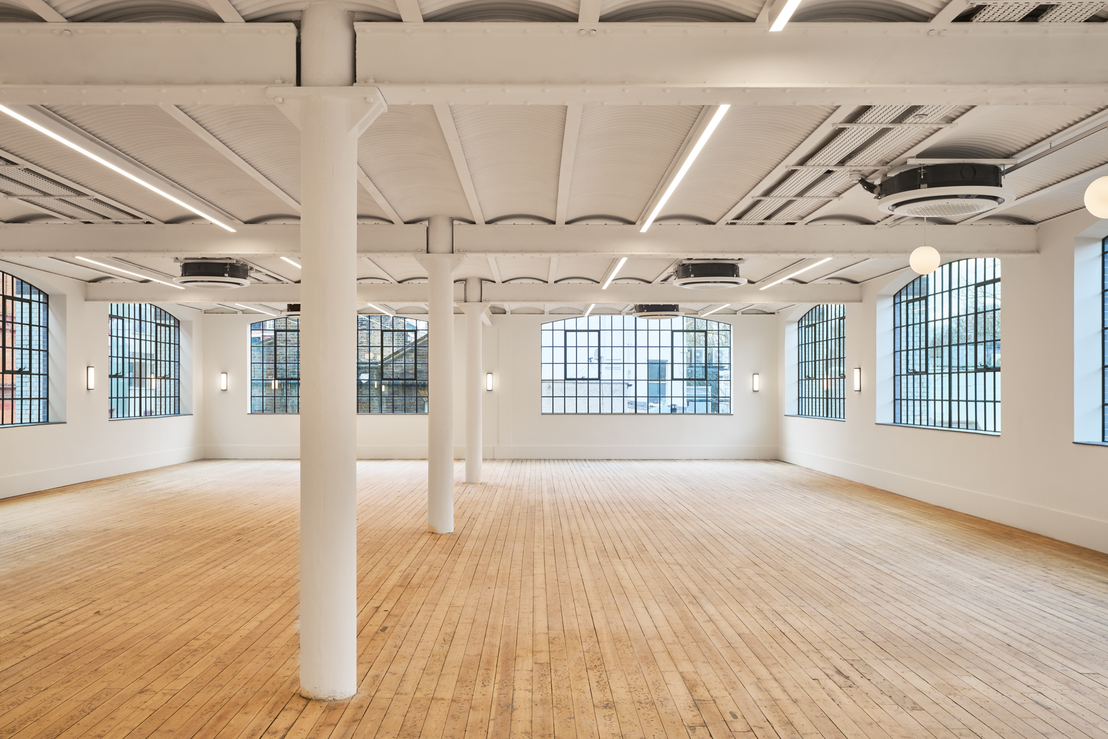 An expansive indoor area at Voysey House, with an arched ceiling and large industrial-style windows. White pillars support the light, wooden, open-plan space.