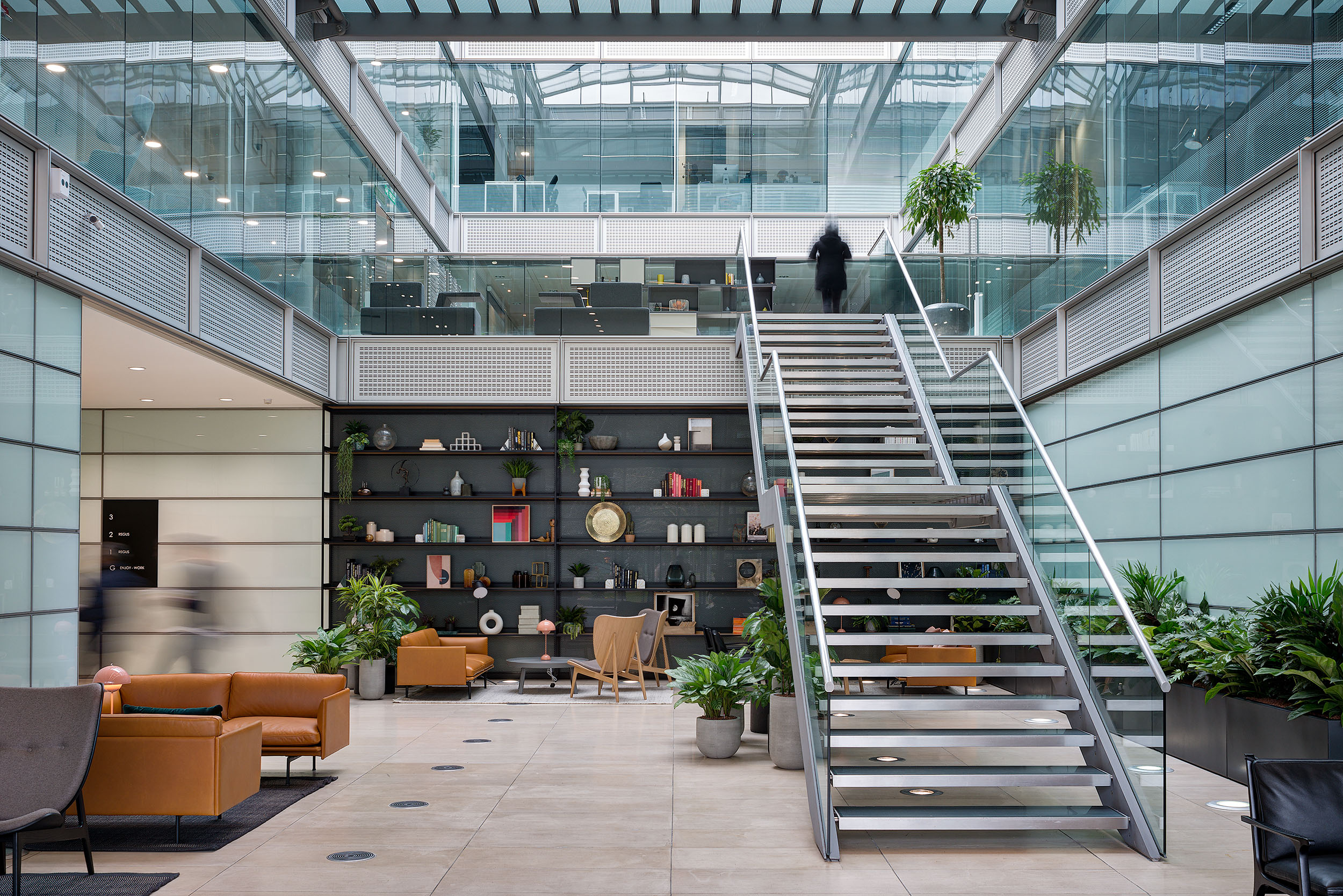 Stylish atrium at Chiswick Business Park Building 3, featuring a central staircase, curated shelving with decorative items, comfortable seating, and lush greenery, all within a bright, modern space with glass partitions and natural light.