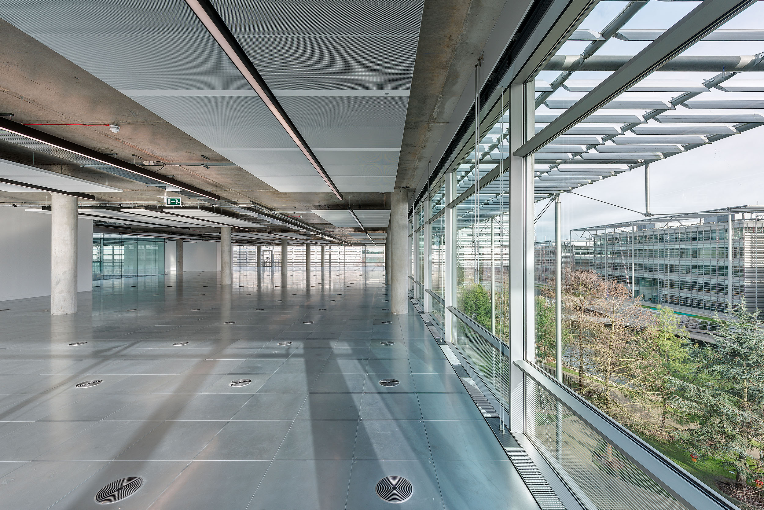 Spacious open-plan area in Chiswick Business Park Building 3, with floor-to-ceiling windows allowing natural light to flow in and views overlooking the surrounding greenery, complemented by an exposed ceiling and polished flooring.