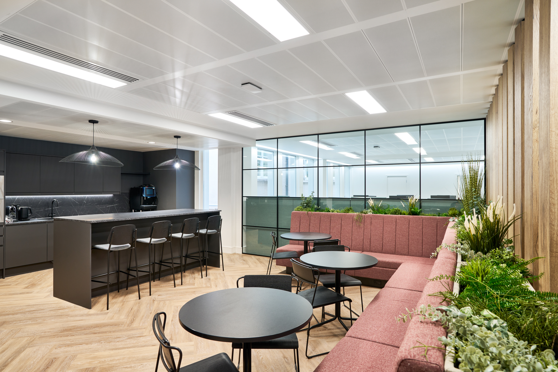 Teapoint and breakout area at Gemserv with a sleek black kitchen island, barstools, pendant lighting, and red banquette seating enhanced with integrated greenery.