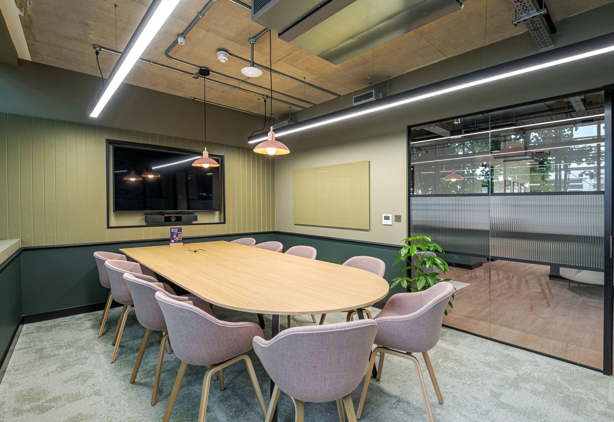 A modern conference room at Work.Life’s Typewriter Building, featuring a large wooden table, pastel chairs, green walls, pendant lighting, and glass partitions.