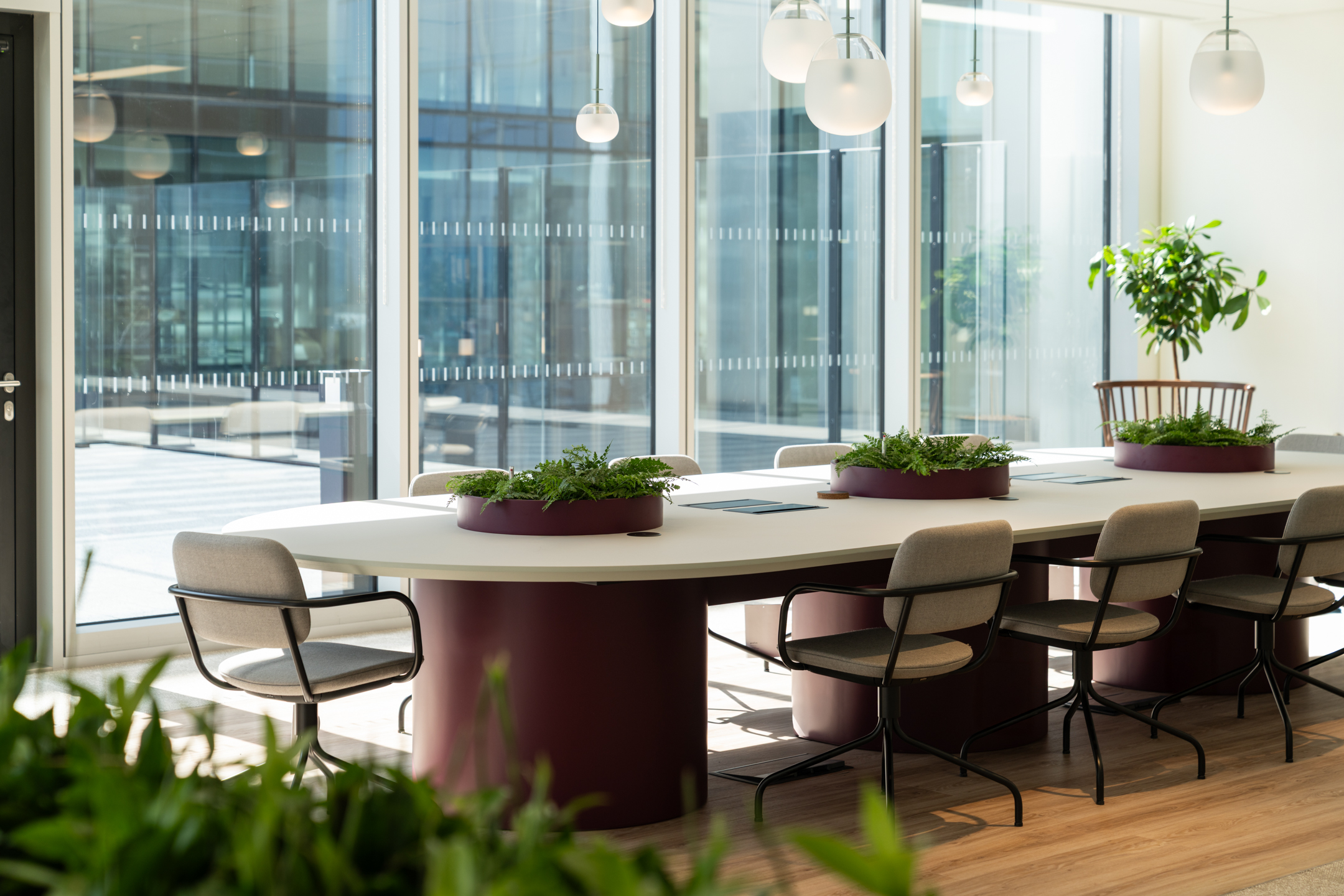 Sunlit workspace at Ark Syndicate with a large central table, built-in planters, and contemporary pendant lighting surrounded by floor-to-ceiling glazing.