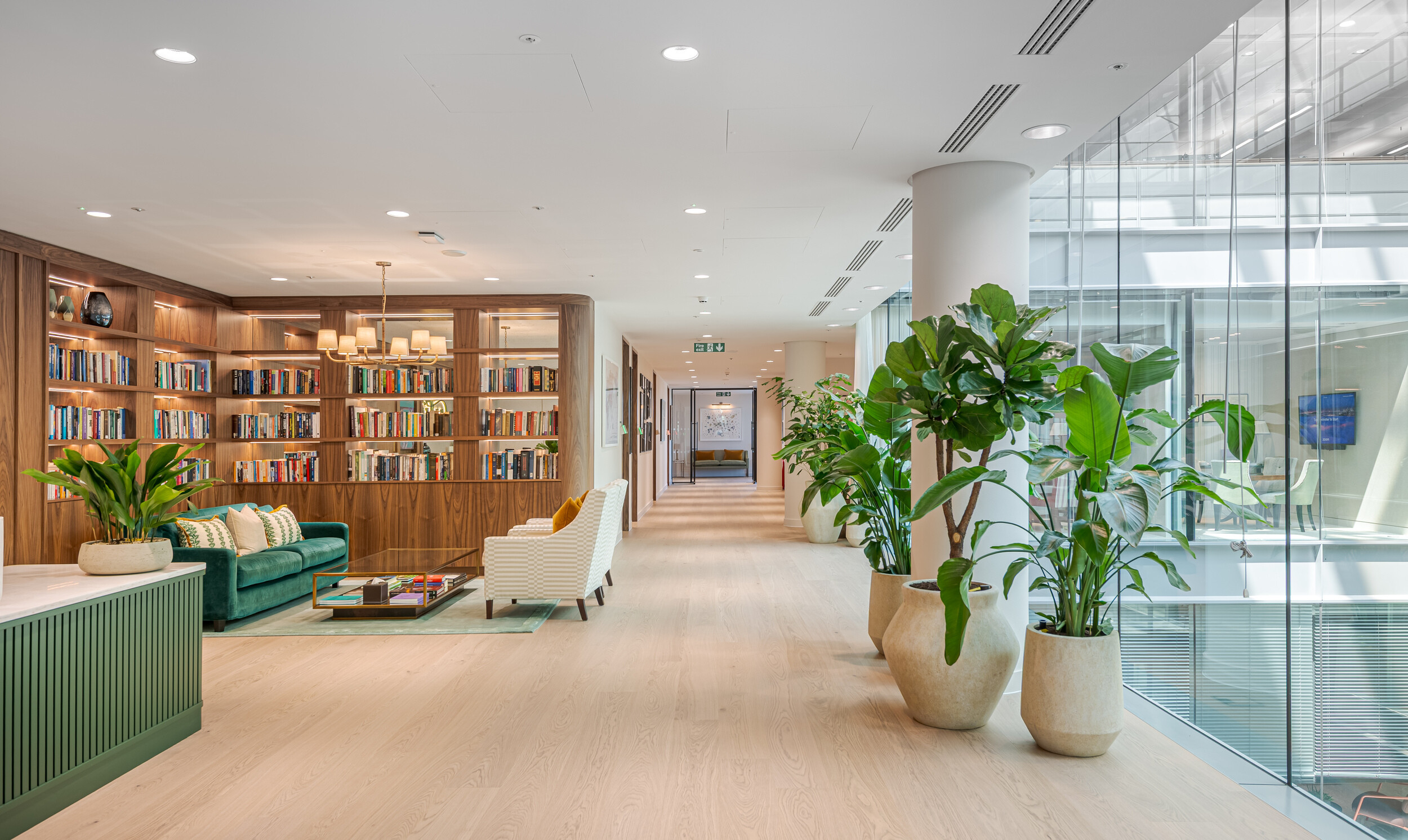 Elegant lounge space within an Investment Management Firm, featuring a library wall with warm timber joinery, velvet seating, and biophilic touches. Large potted plants line the corridor beside full-height glazing, bringing natural light into the fit out. Designed to feel welcoming and refined.