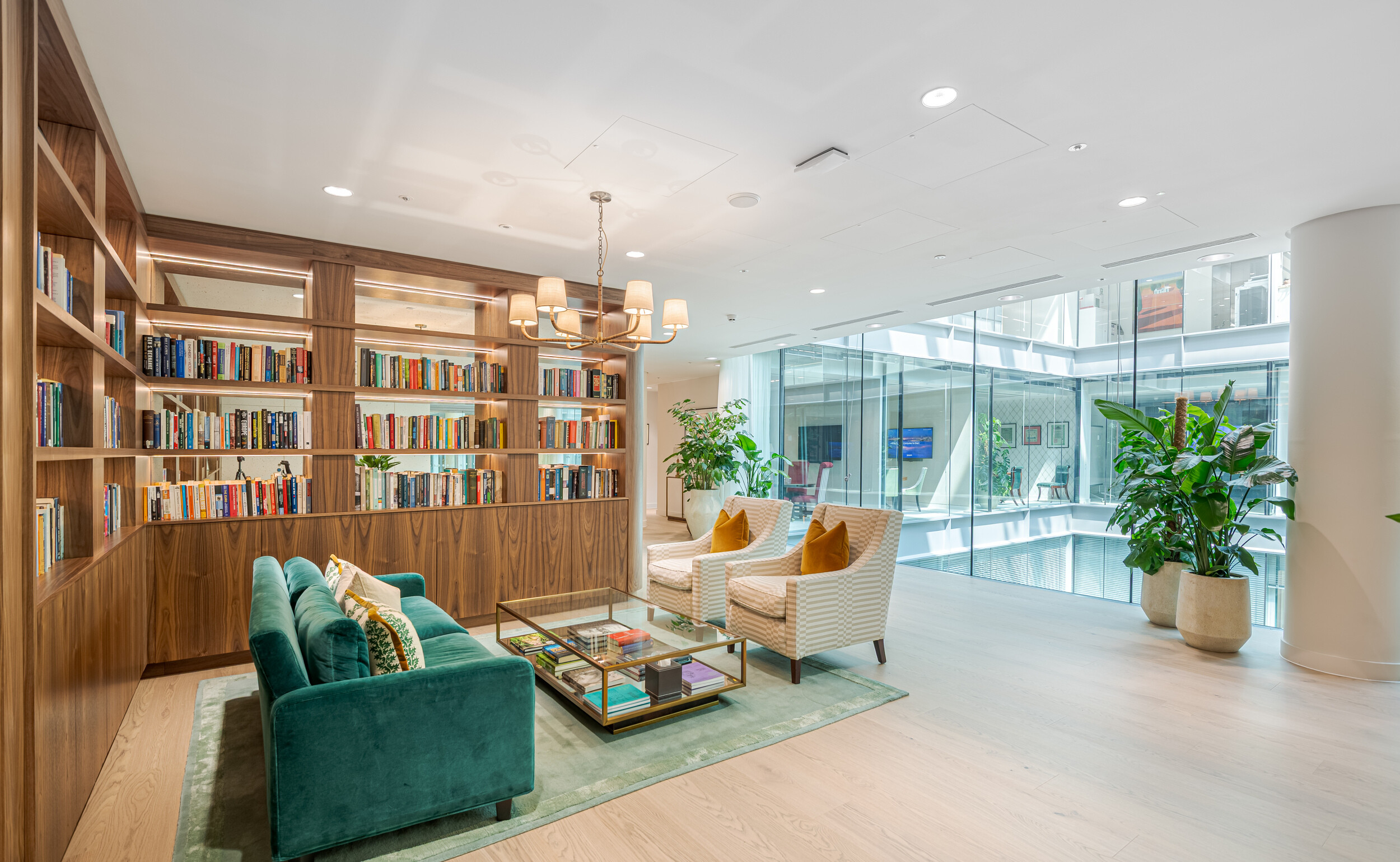 Bright, relaxed lounge in an Investment Management Firm, showcasing a built-in timber library, green velvet sofa, and striped armchairs with mustard cushions. A large glass coffee table sits on a soft rug beside expansive internal glazing. The fit out prioritises comfort and clarity in a professional setting.
