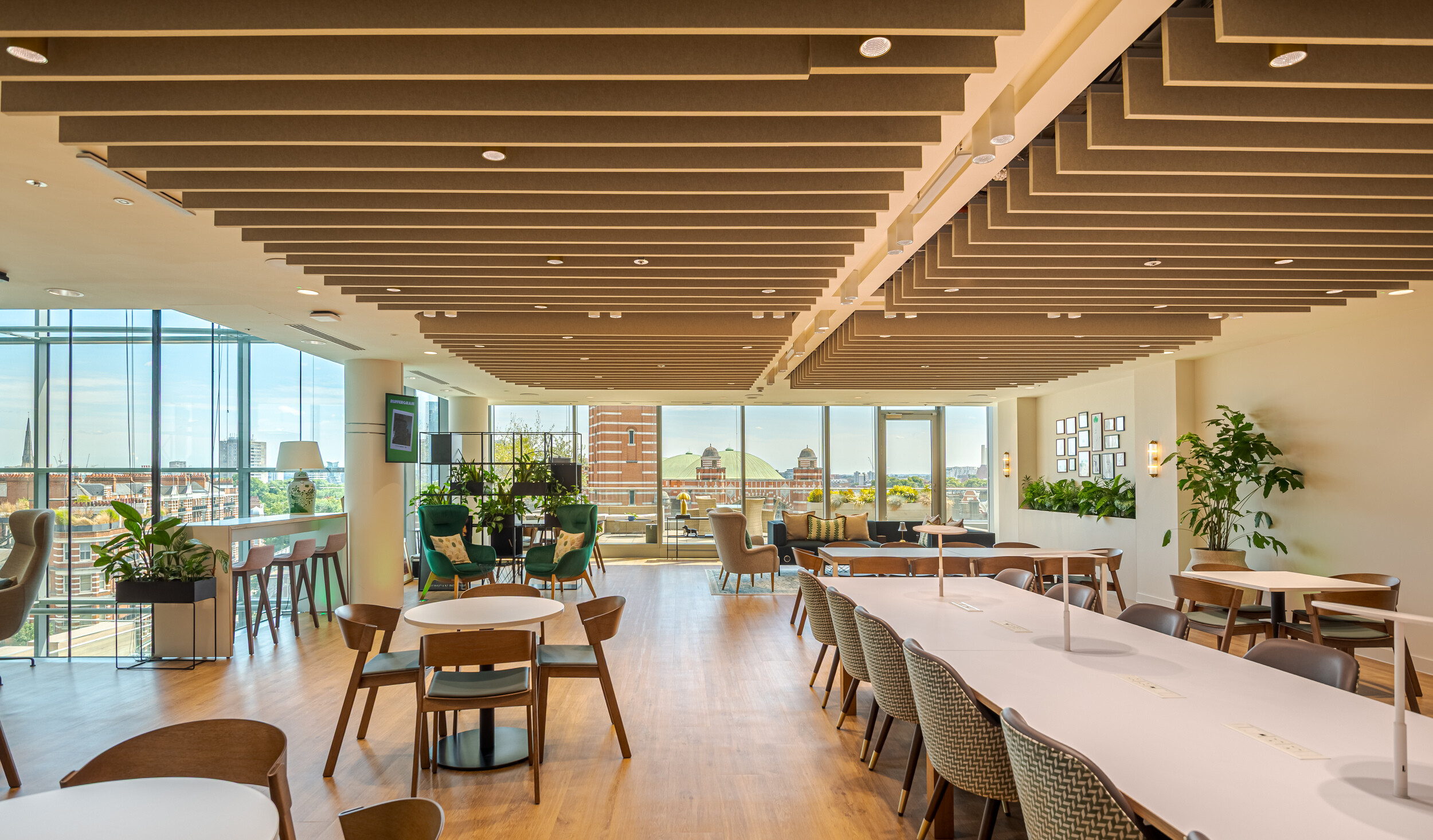 Flexible breakout and café-style space within an Investment Management Firm, with acoustic ceiling baffles, diverse seating, and panoramic city views. A mix of dining tables, lounge chairs, and soft planting create a relaxed yet functional environment. Floor-to-ceiling glazing floods the area with natural light.