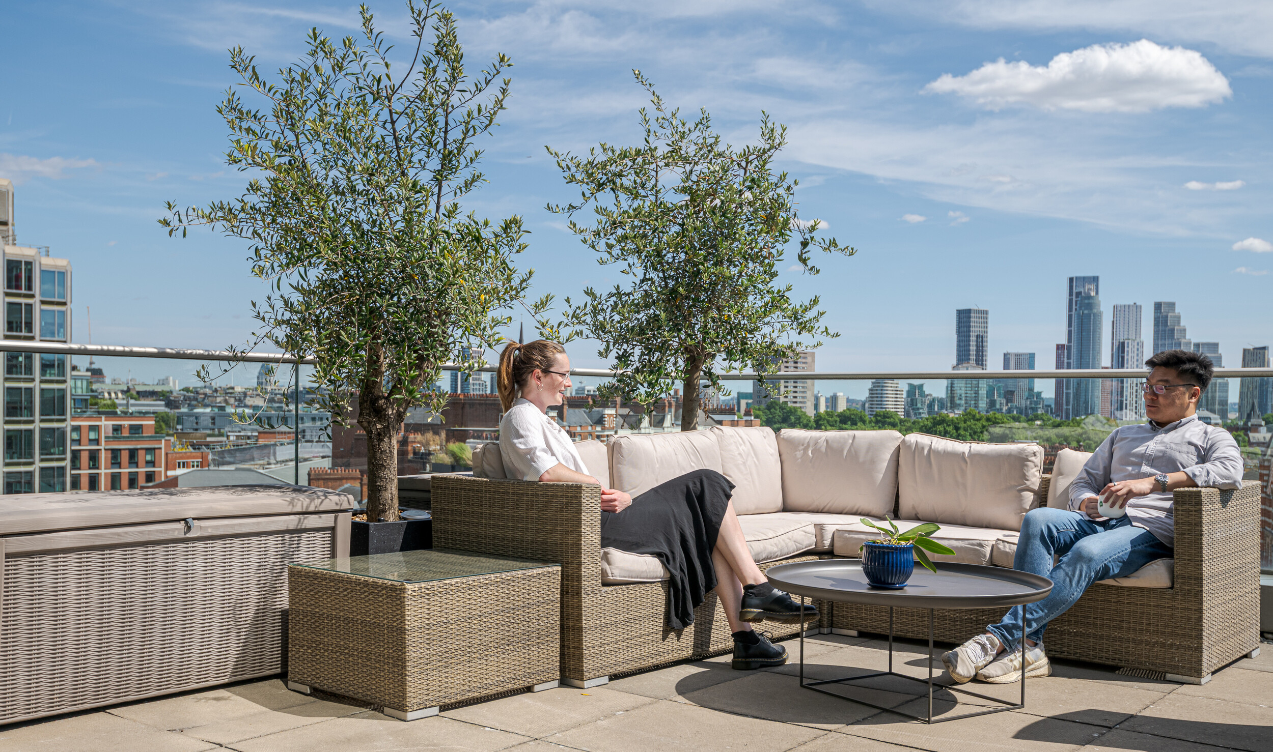 Rooftop terrace at an Investment Management Firm, with rattan-style seating, potted olive trees, and panoramic views across the London skyline. Two colleagues enjoy coffee and conversation in the sunshine. The outdoor space adds a calm, social element to the workplace.