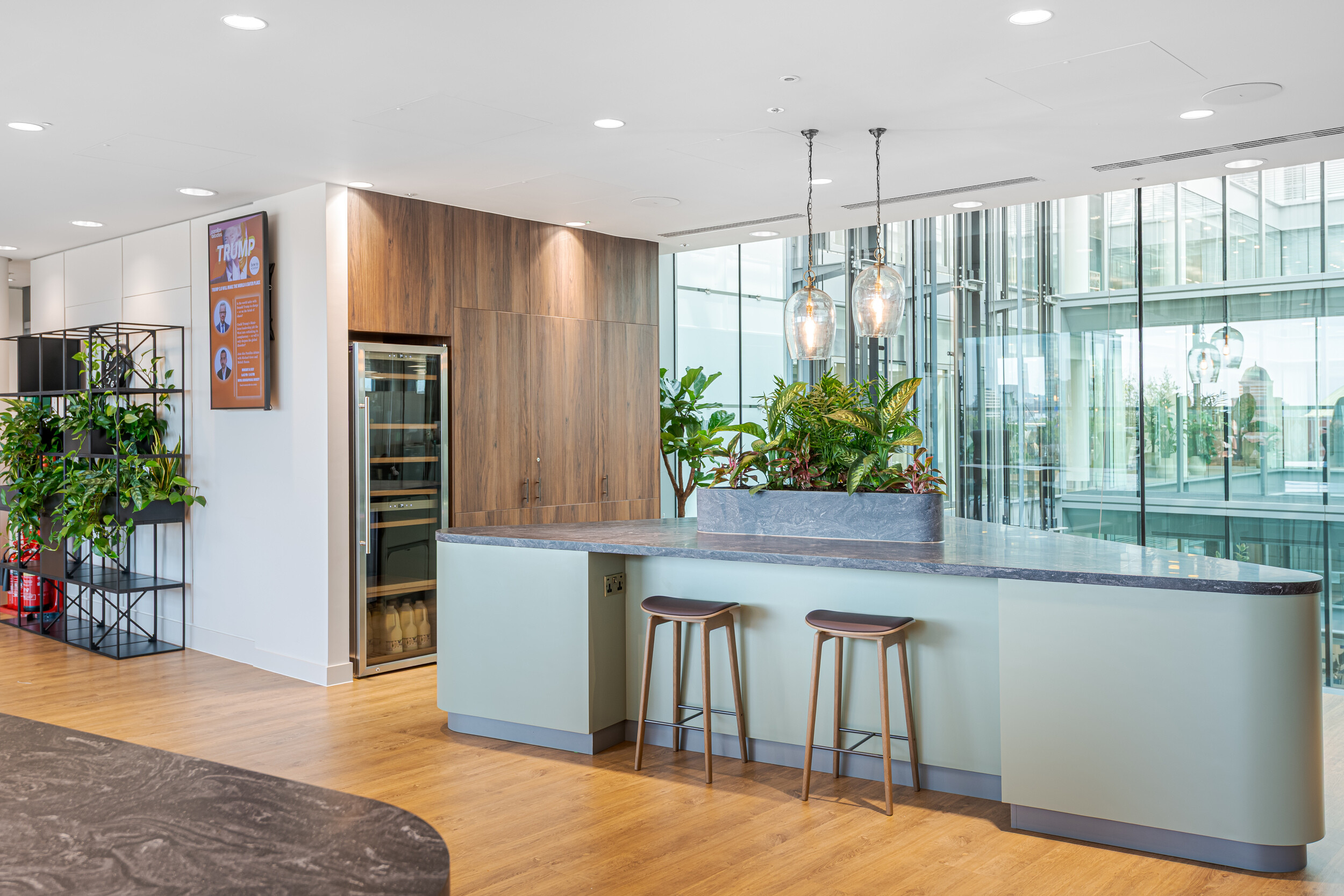 Contemporary tea point in an Investment Management Firm, with curved island seating, pendant lights, and timber cabinetry. A central planter adds greenery beneath a glazed atrium, enhancing the biophilic design. Neutral tones and modern finishes create a calm and inviting break space.