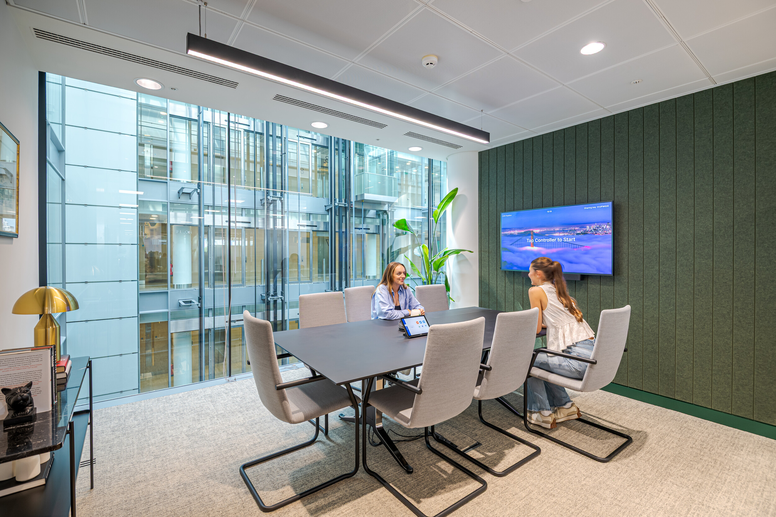 Small meeting room in an Investment Management Firm, featuring a sleek black table, grey chairs, and a green acoustic-panelled wall. Two colleagues hold a discussion beside a floor-to-ceiling glazed atrium. Integrated tech and natural light support a modern, focused environment.