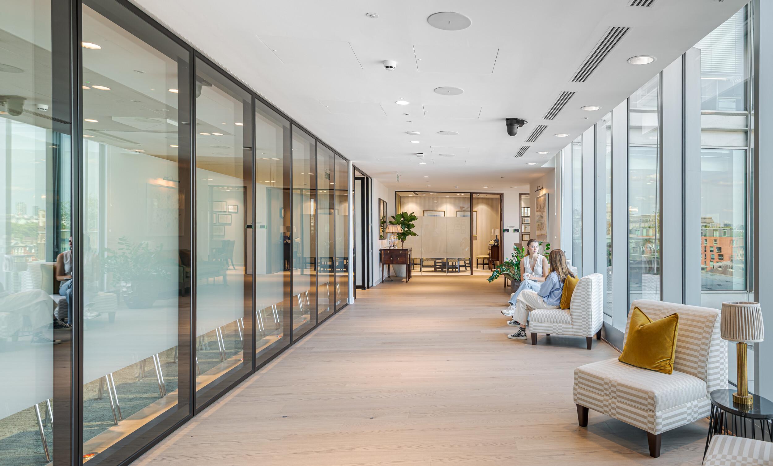 Bright corridor in an Investment Management Firm, lined with glazed meeting rooms on one side and soft seating overlooking city views on the other. Two colleagues chat informally in striped armchairs with mustard cushions. Timber floors, biophilia and elegant lighting complete the relaxed setting.