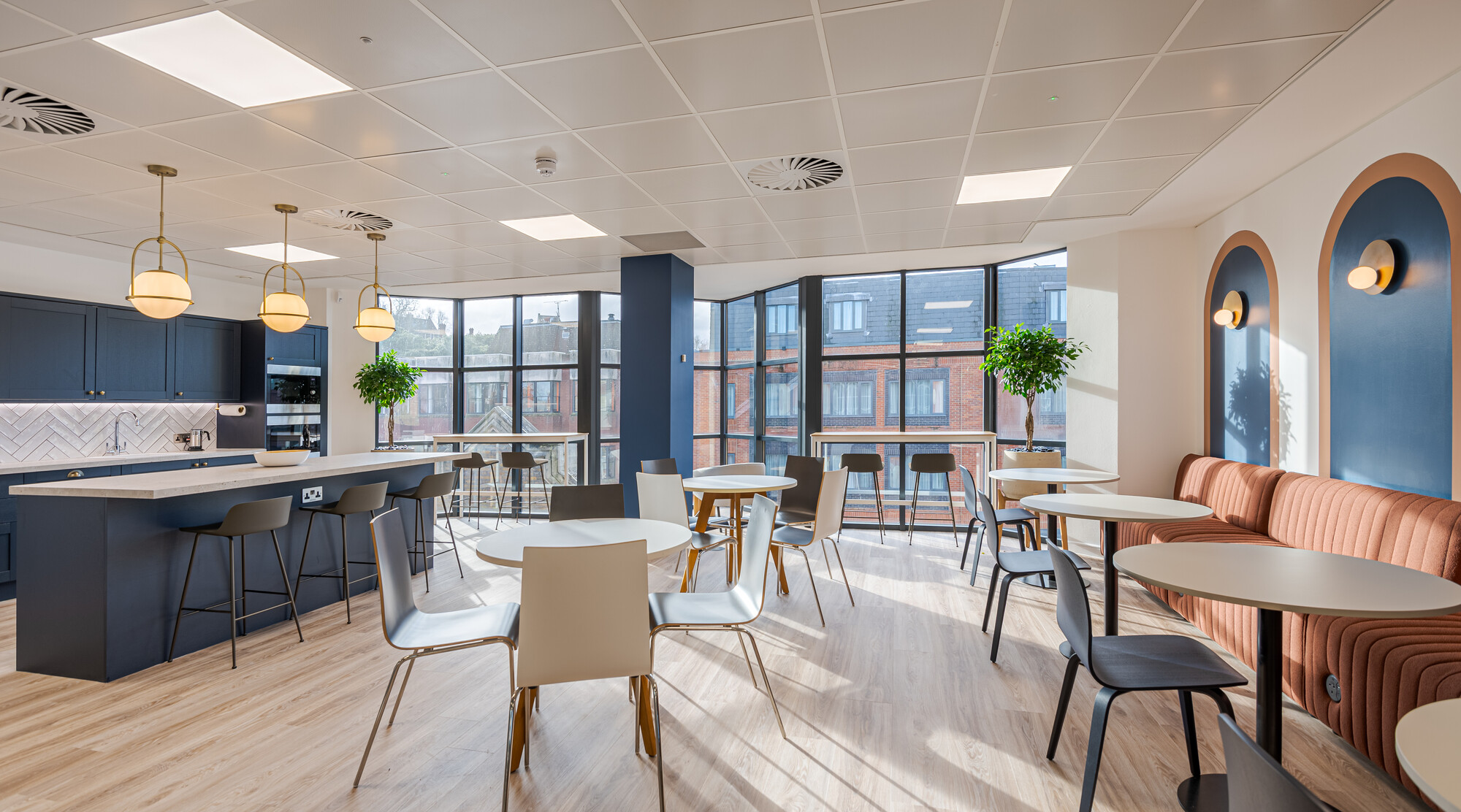 Accelya office designed by Two, featuring a kitchen and dining area with a navy blue island, herringbone-tiled backsplash, and brass pendant lights.