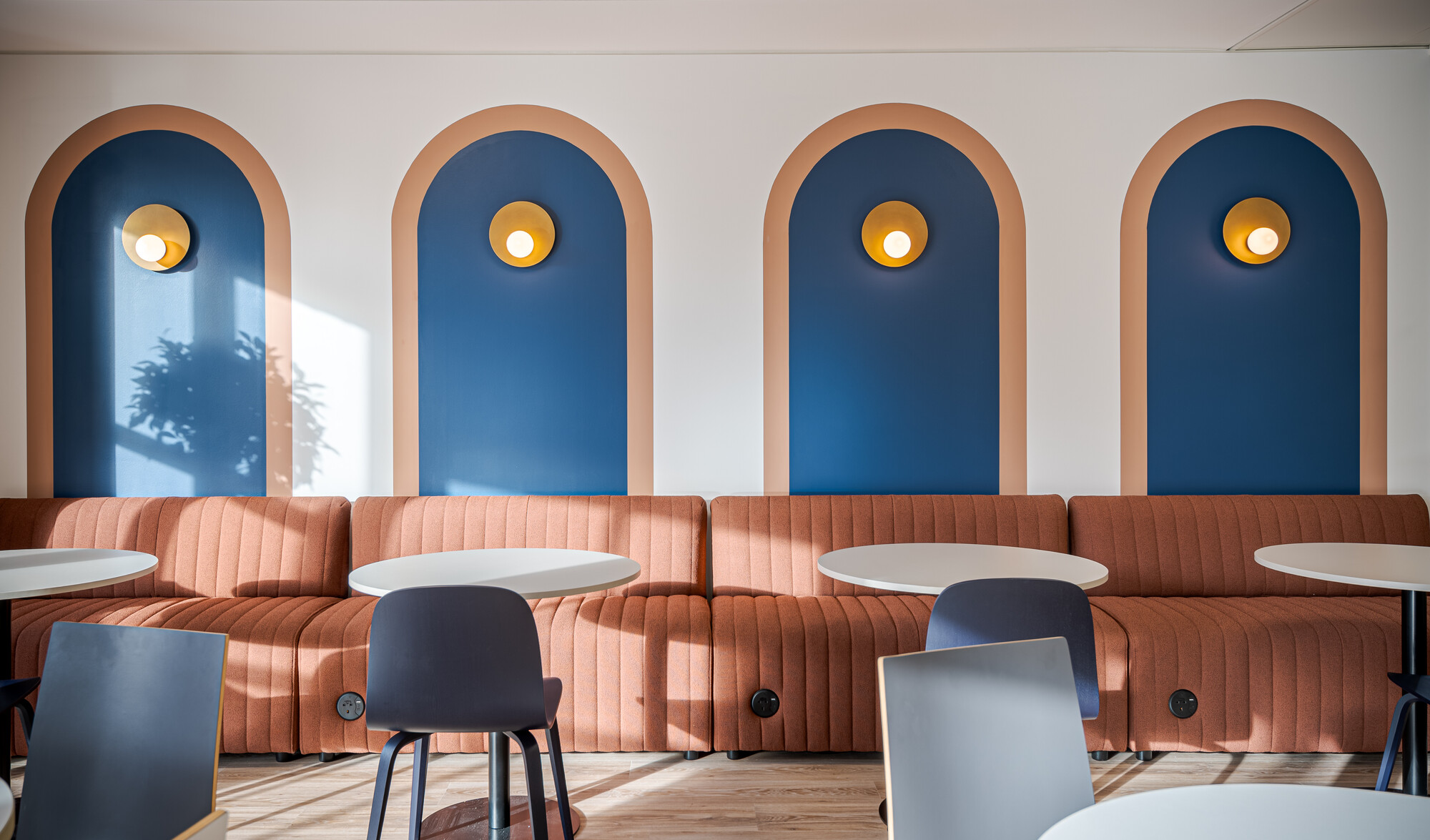 Accelya office designed by Two, showing a seating area with a rust-coloured ribbed banquette, round white tables, and navy blue chairs.