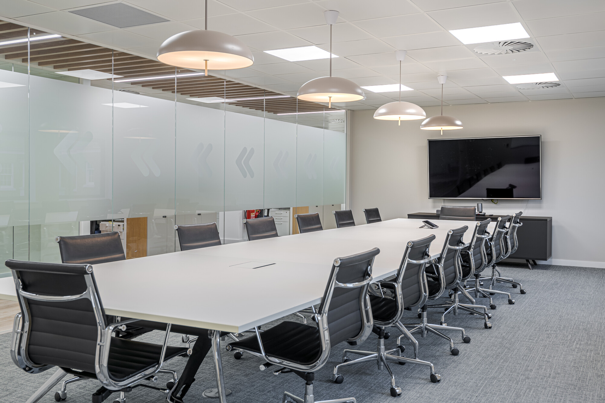 Large meeting room in Accelya's office by Two, with a long white conference table surrounded by black leather chairs on casters. Frosted glass walls with arrow motifs separate the space, and pendant lighting hangs overhead.