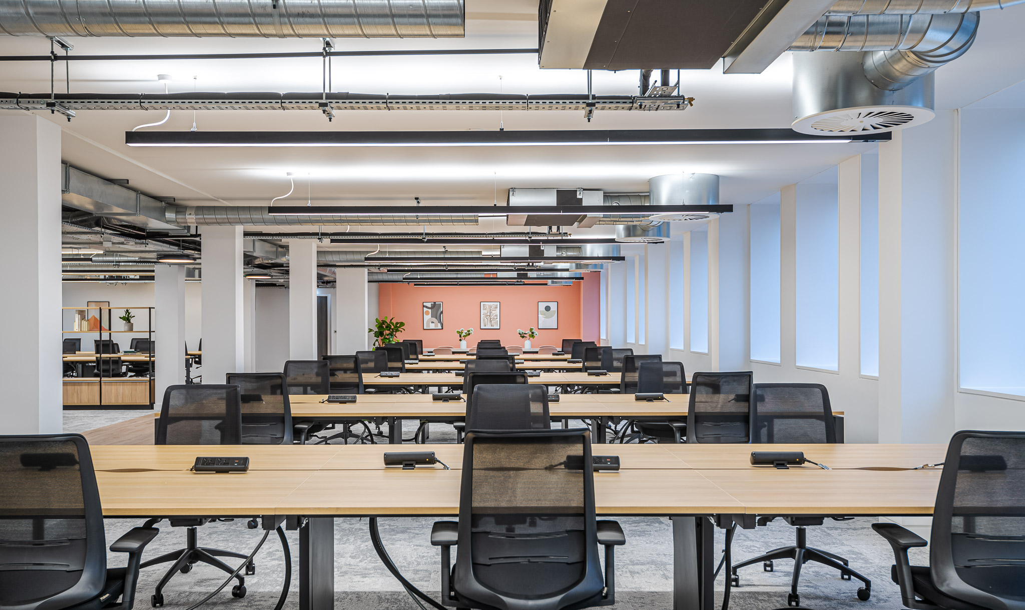 Desking area within the Asset Management Firm workspace, featuring long workbenches, ergonomic chairs, and a soft coral accent wall to add warmth to the structured layout.