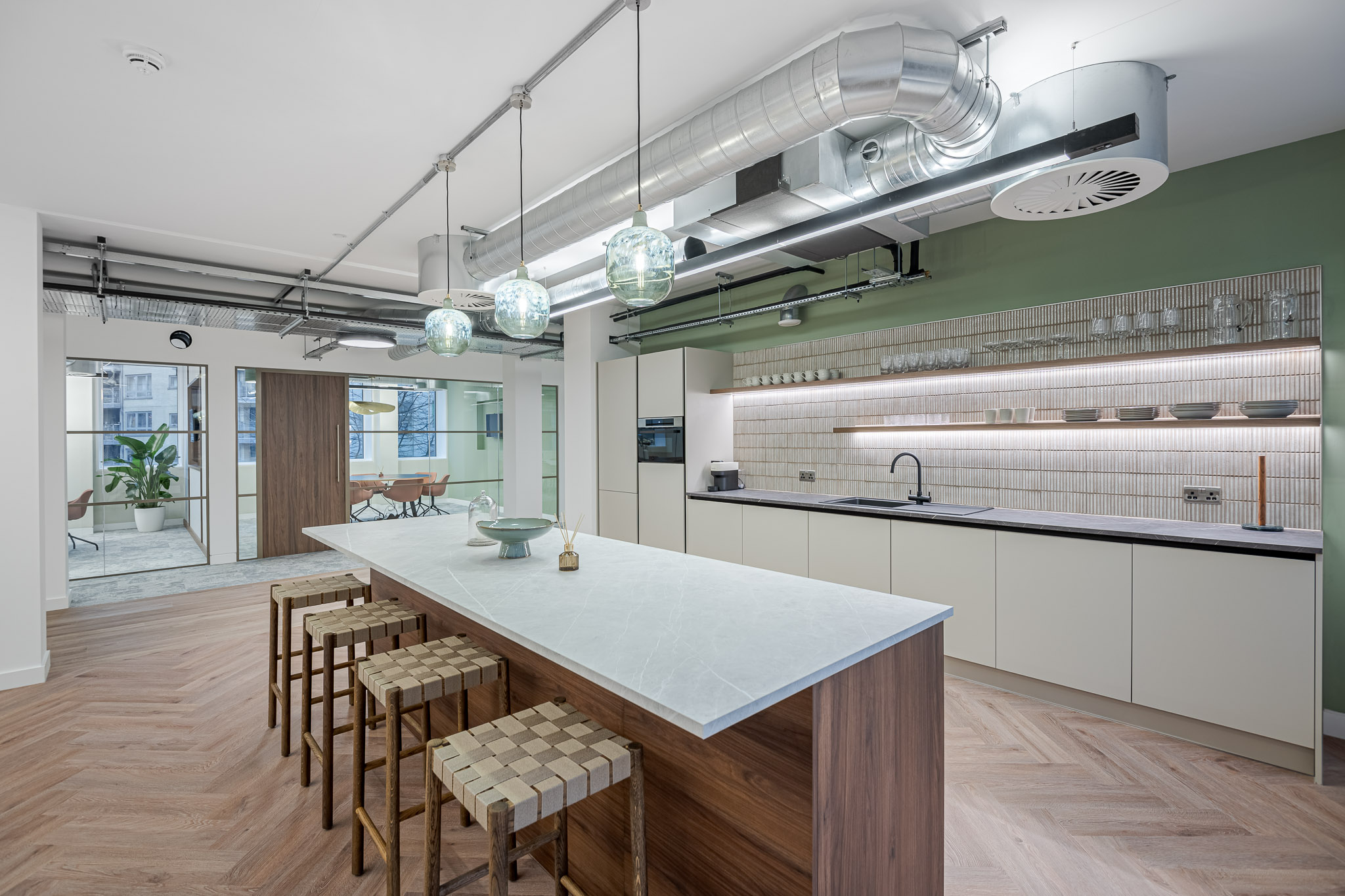 Modern teapoint in the Asset Management Firm's Tenant-Ready™ space, featuring a marble-topped island, green accent wall, open shelving, and pendant lights with a view into surrounding meeting areas.