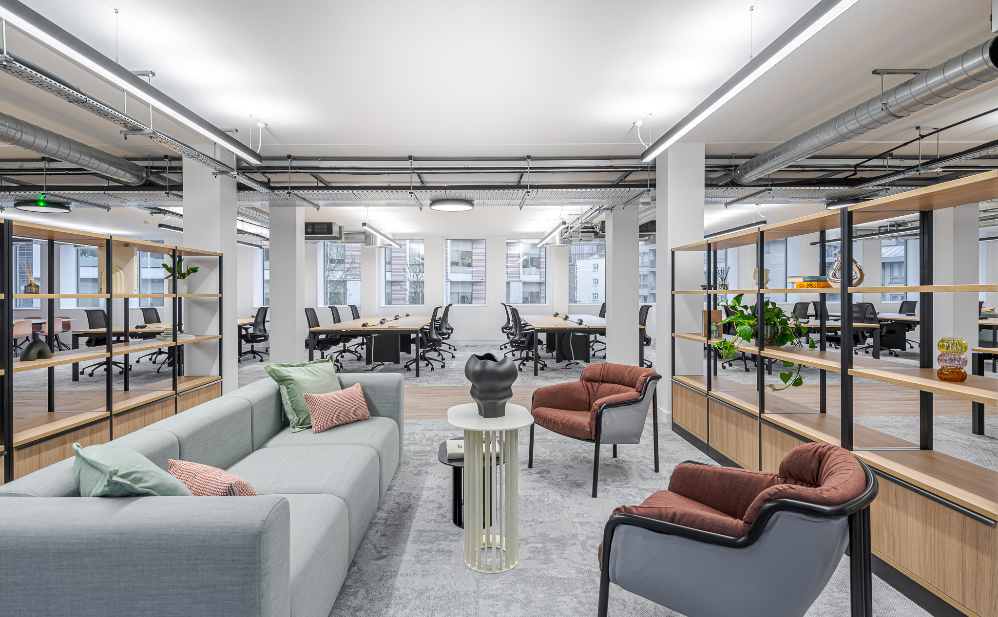 Tenant-Ready™ Breakout seating area in the Asset Management Firms Tenant-Ready™ workspace, framed by open shelving and surrounded by open-plan desks, blending soft furnishings with an industrial ceiling design.