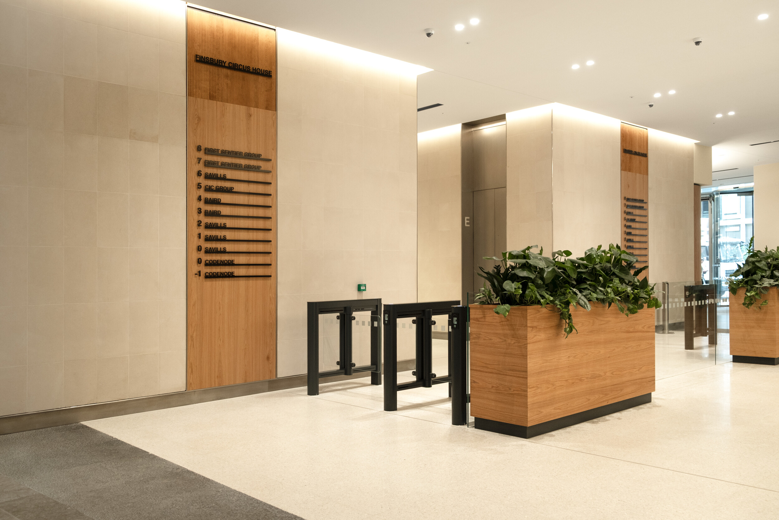 Ground floor lobby at Finsbury Circus House with sleek lift entrances, wood-accented tenant directory, secure entry gates, and large planters with greenery.
