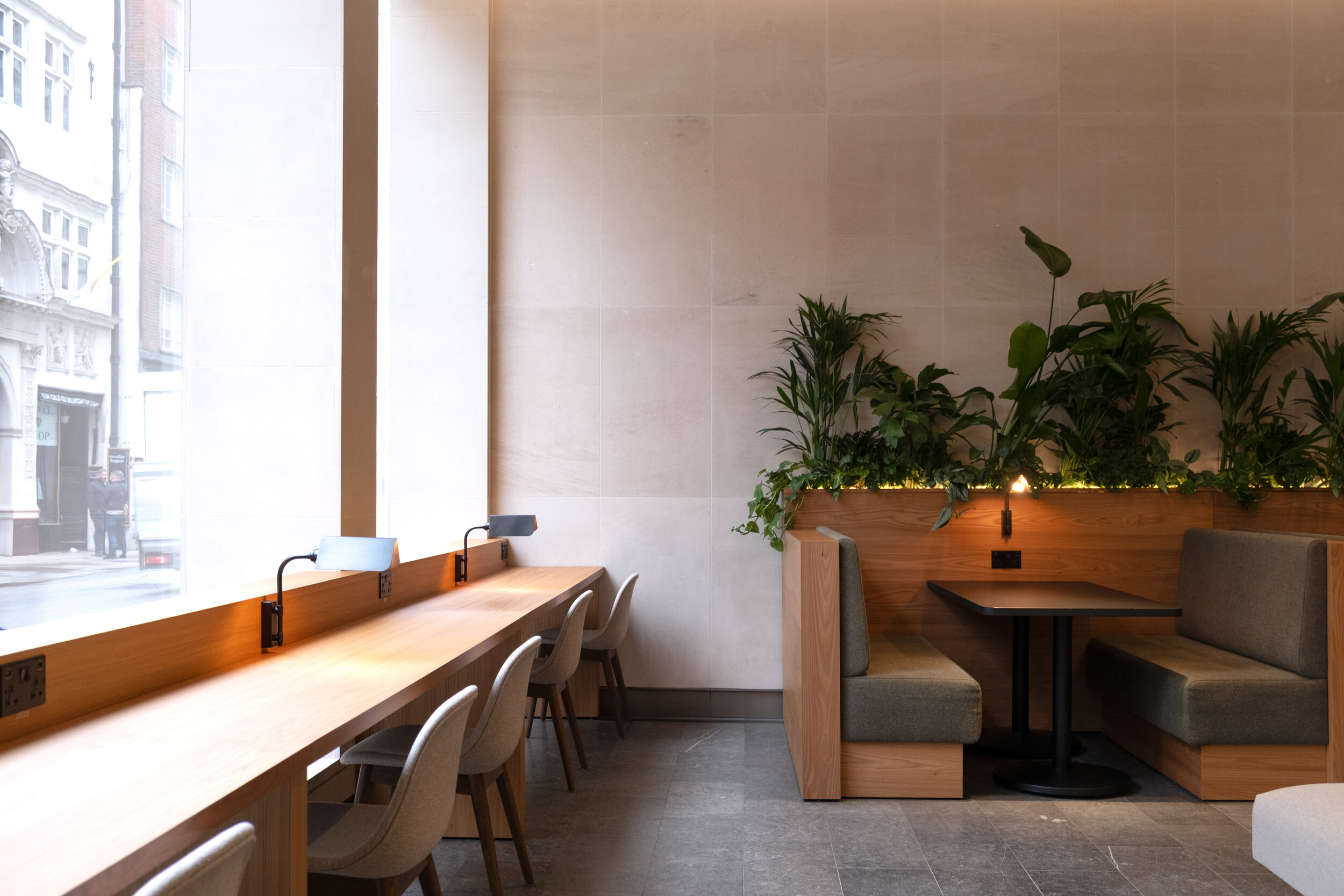 Cosy workspace at Finsbury Circus House with a long wooden desk by the window, upholstered chairs, and booth seating framed by lush green plants.