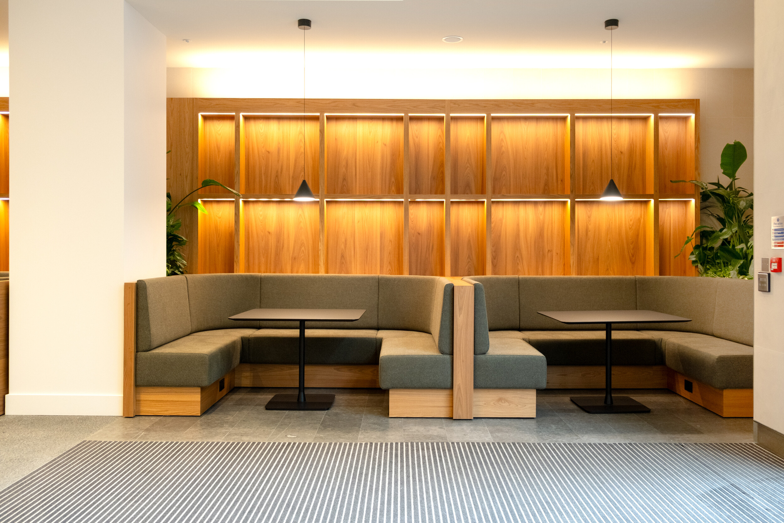 Stylish booth seating area at Finsbury Circus House with green upholstered benches, black tables, and a warm wood-panelled feature wall with pendant lighting.