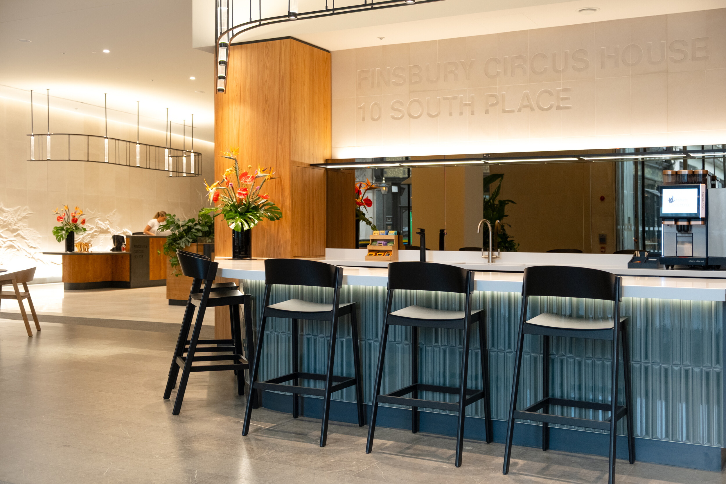 Modern bar-style seating area at Finsbury Circus House with high black chairs, textured teal panelling, and vibrant floral arrangements against a wood and stone backdrop.