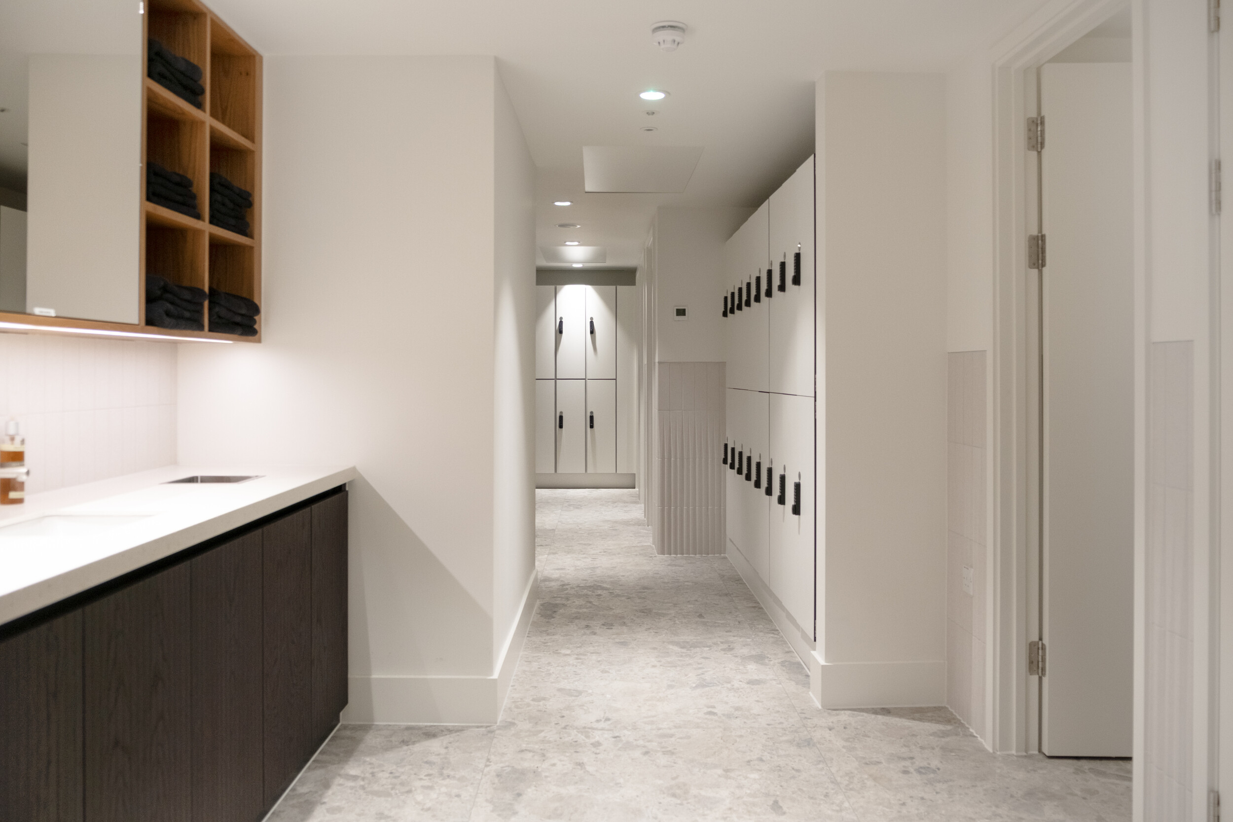 Changing room at Finsbury Circus House with sleek white lockers, stone flooring, dark wood cabinetry, and neatly stacked towels.