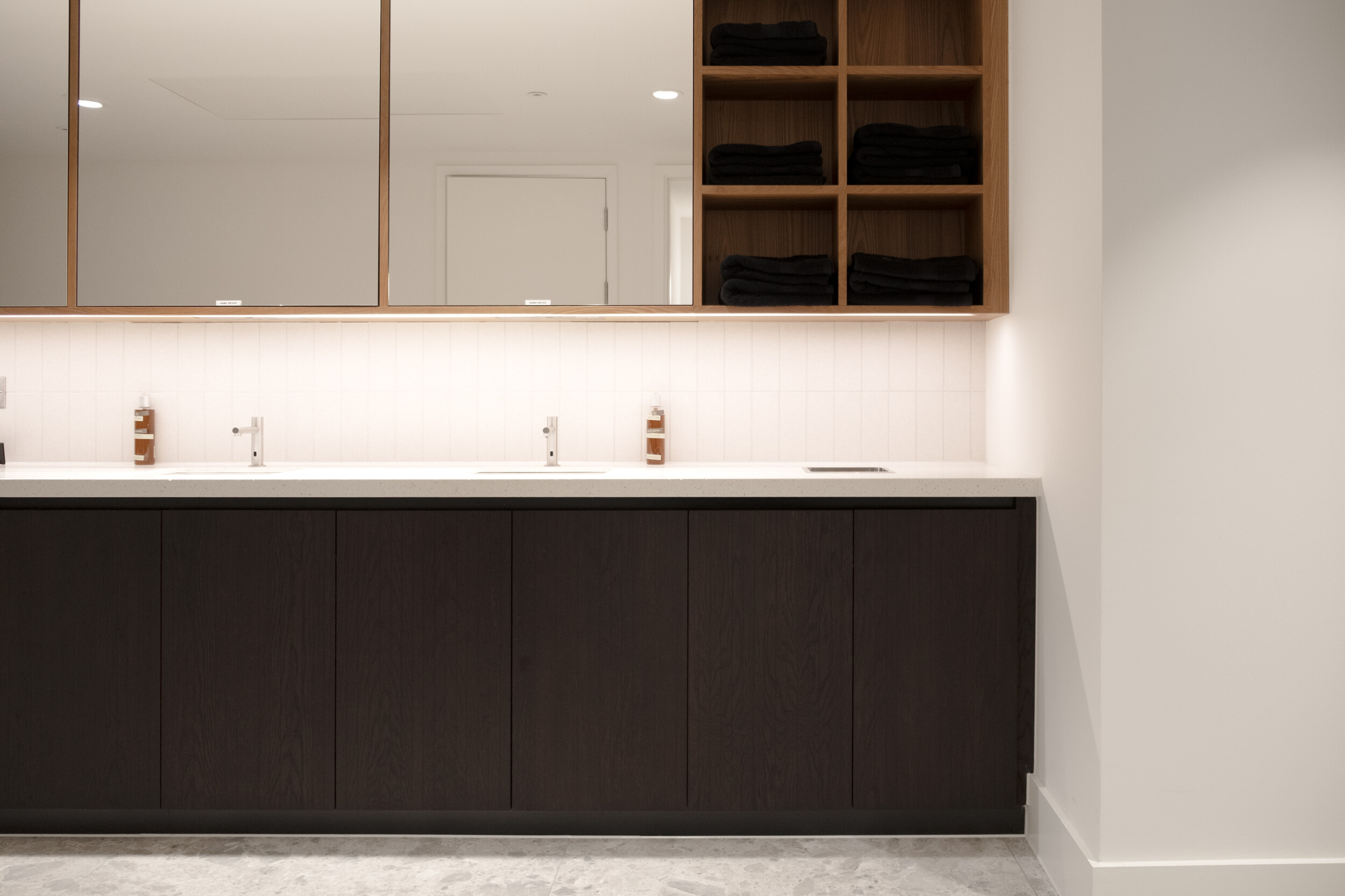 Washroom area at Finsbury Circus House with double sinks, dark wood cabinetry, large mirrors, and open shelving stocked with neatly folded black towels.
