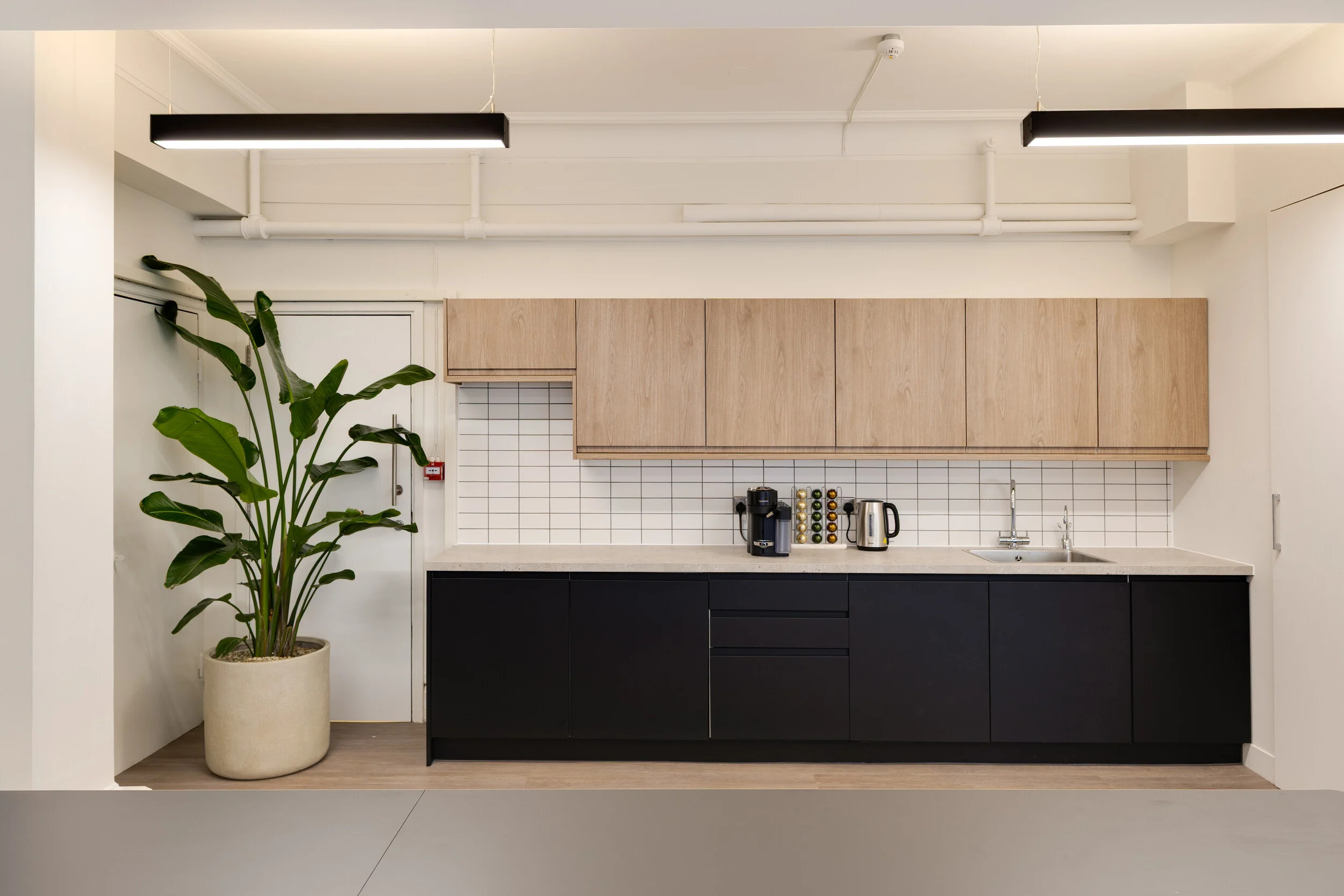 Modern teapoint for InPost with black lower cabinets, light wood wall units, coffee machine, kettle, and a large indoor plant.