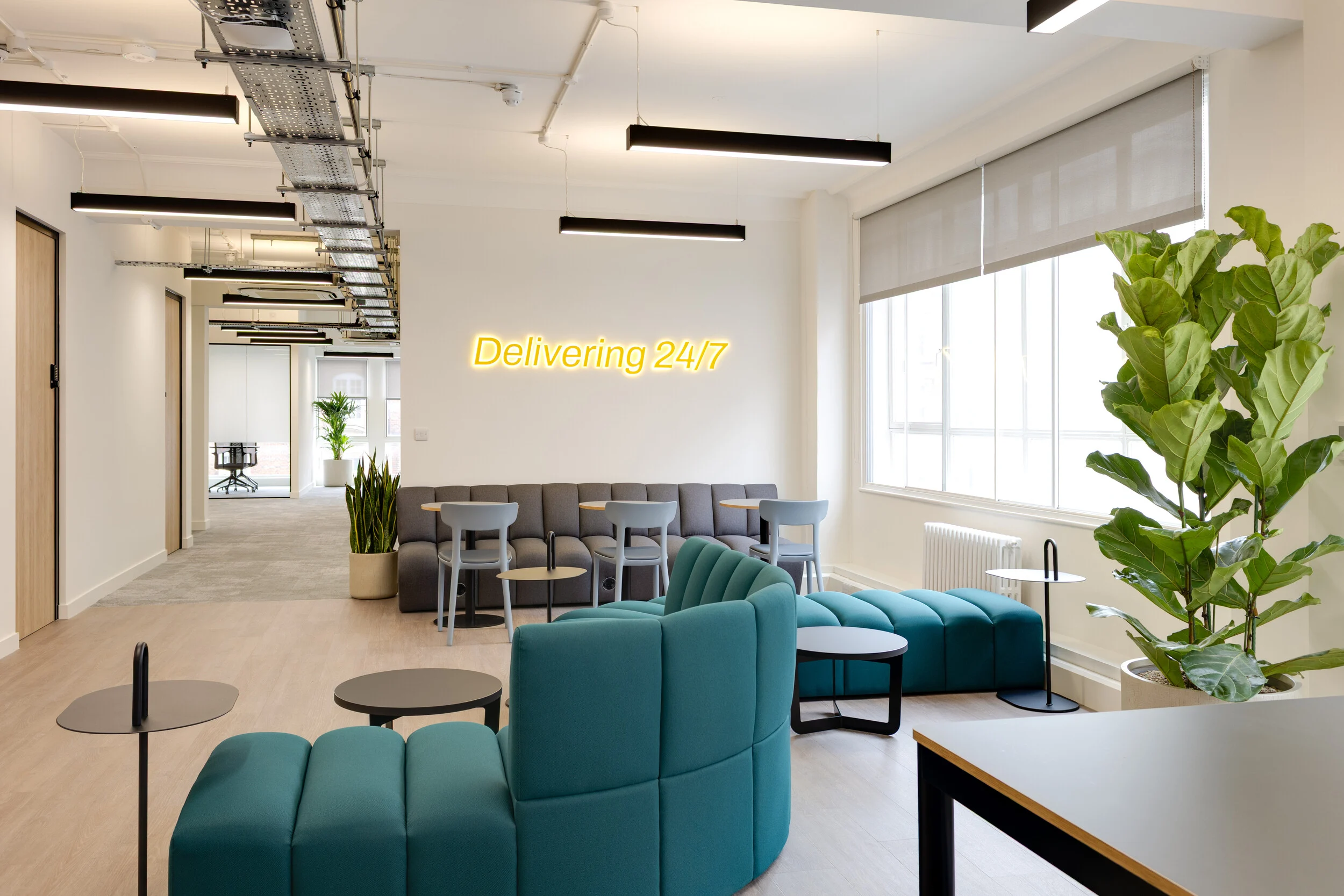 InPost breakout space with teal modular seating, grey tables and chairs, potted greenery, and a glowing yellow 'Delivering 24/7' sign by the window.