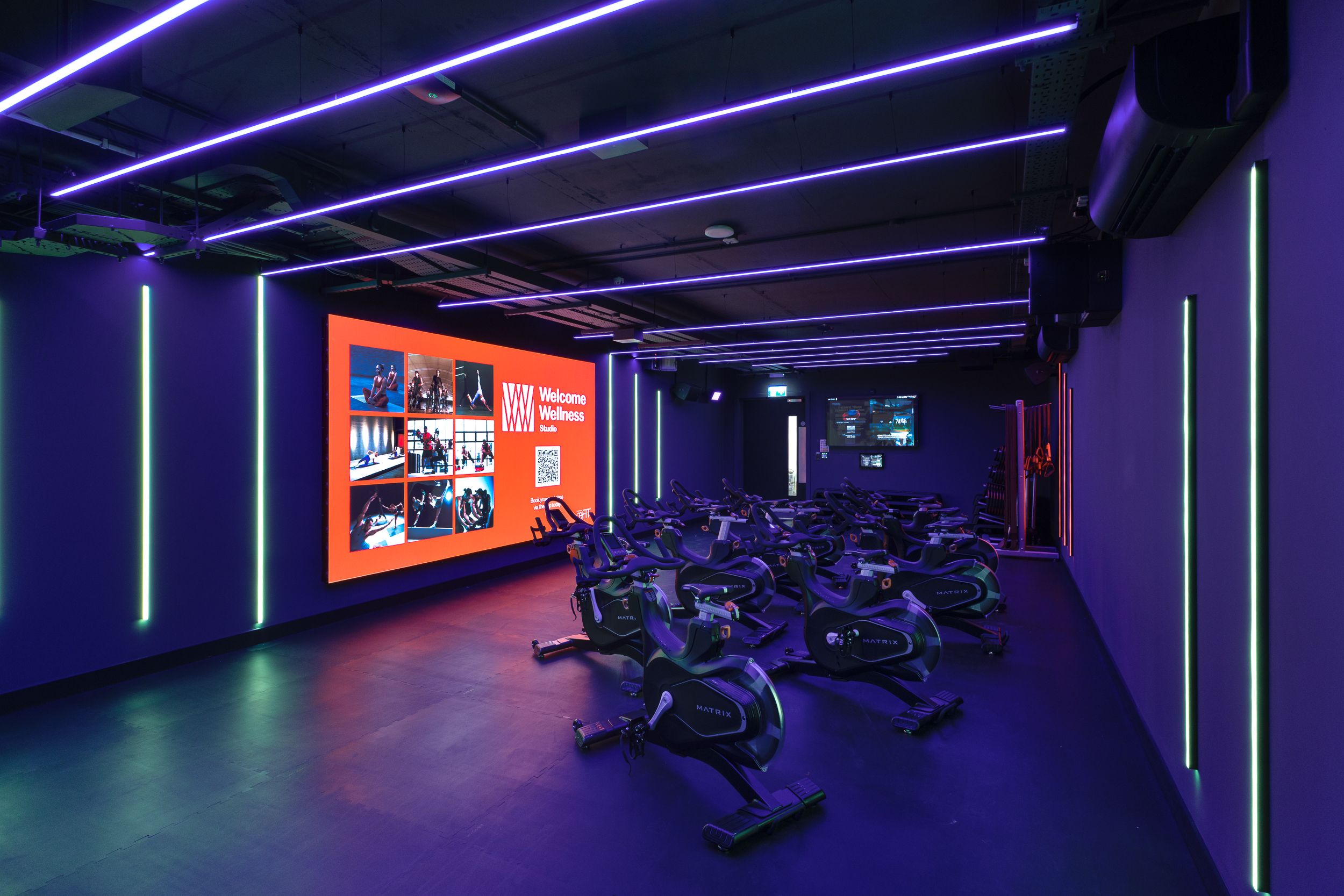 Indoor cycling studio at reFIT The Welcome Building with rows of Matrix bikes, illuminated by purple and green LED lighting and a large digital wellness display.