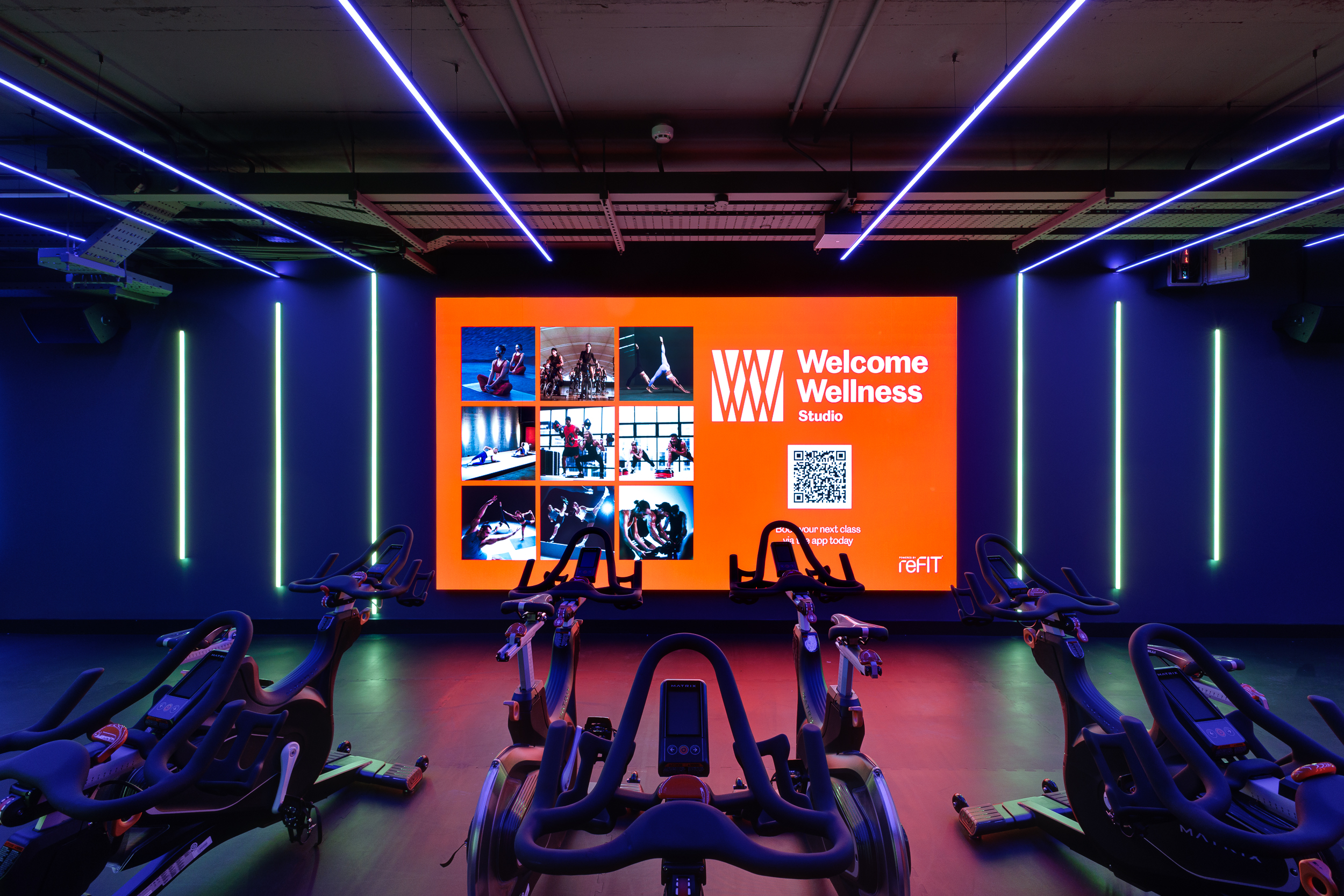 Front view of the cycling studio at reFIT The Welcome Building, showcasing Matrix bikes and a large digital Welcome Wellness screen illuminated by blue and purple LED lights.