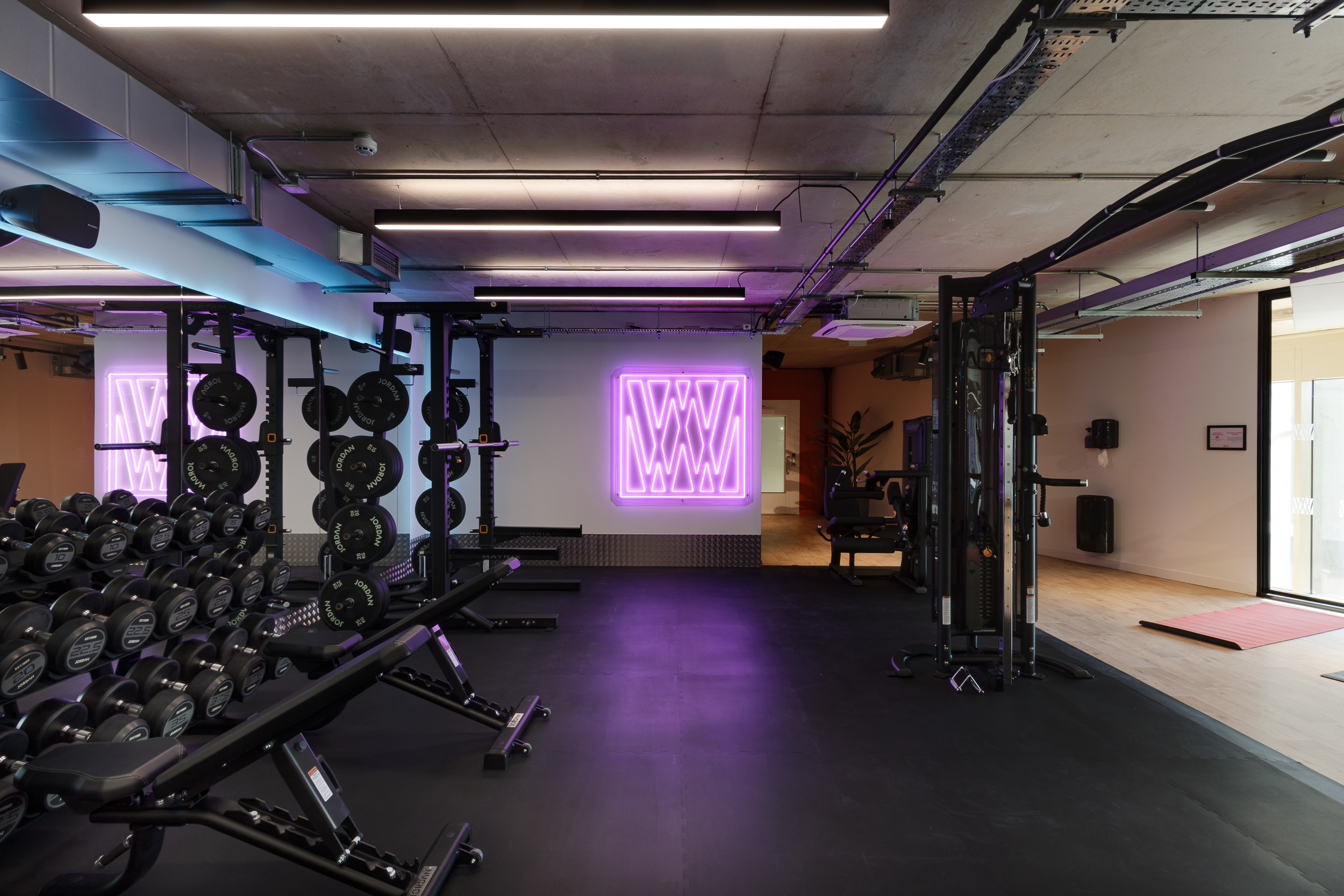 Modern gym at reFIT The Welcome Building featuring free weights, squat racks, and a bright purple Welcome Wellness neon sign adding a vibrant touch to the workout space.