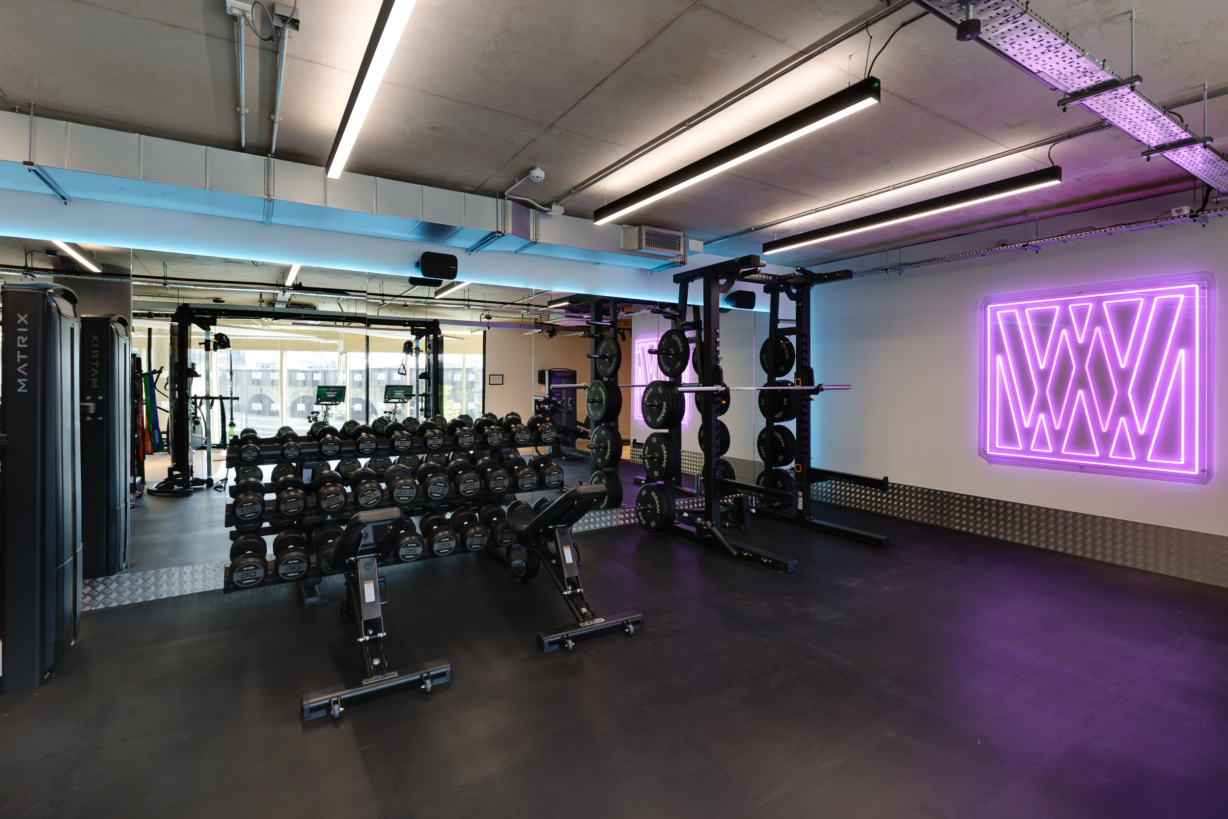 Gym space at reFIT The Welcome Building with free weights, squat racks, and strength equipment, highlighted by a glowing purple Welcome Wellness neon sign.
