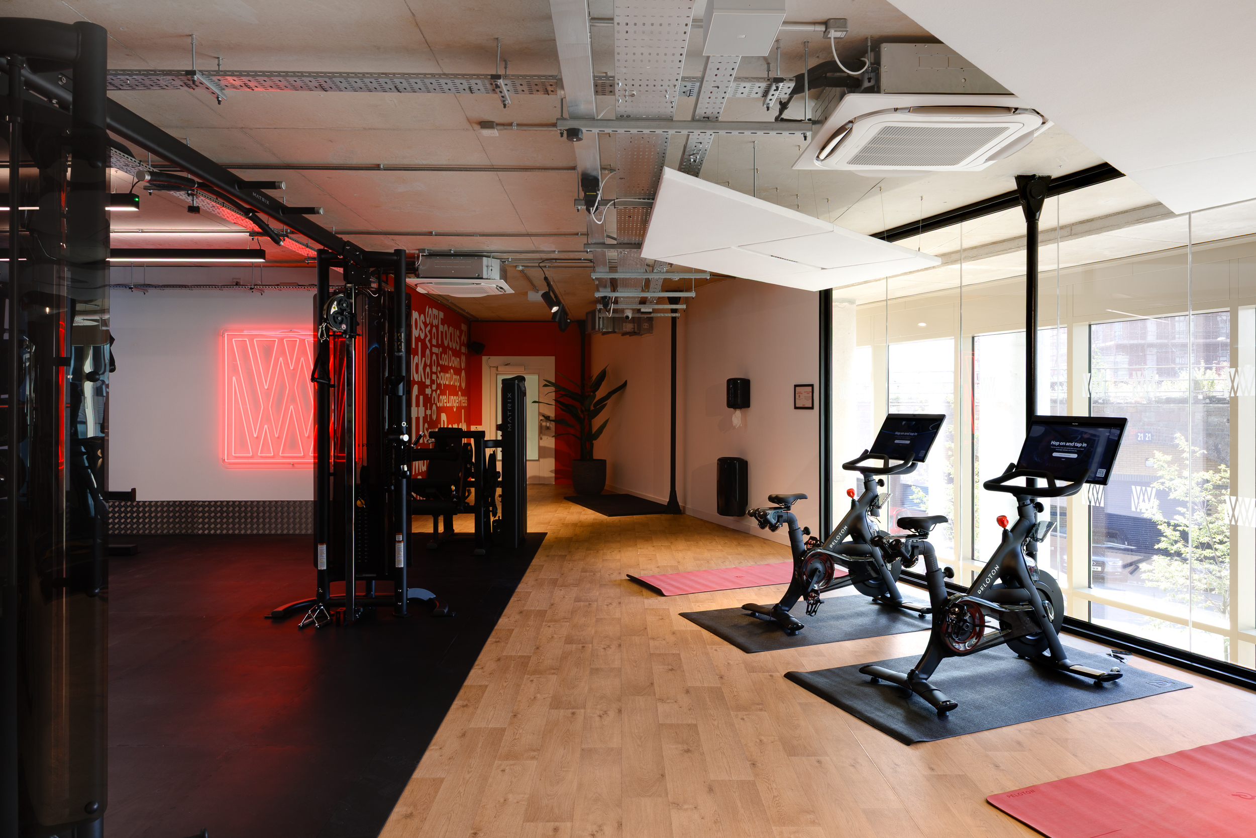 Bright gym area at reFIT The Welcome Building featuring indoor bikes by the windows, yoga mats, and a red Welcome Wellness neon sign in the background.